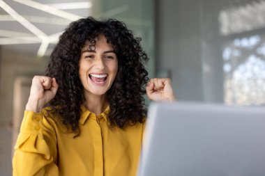 Neşeli bir kadın heyecan içinde yumruklarını sıkar, dizüstü bilgisayar ekranına bakarken mutluluğunu ifade eder. O sarı bir gömlek giyiyor..