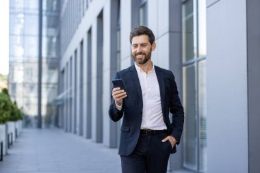 Takım elbiseli gülümseyen bir işadamı dışarıda telefonuna bakıyor. Etrafı modern bir ofis binasıyla çevrili. Adam telefonu elinde tutuyor..