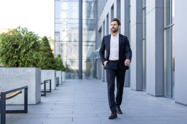 İyi giyimli bir işadamı modern bir şehir ortamında güvenle yürüyor, profesyonel hırsları yayıyor. Koyu renk bir takım giyiyor..