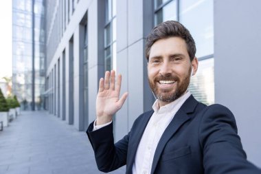 Gülümseyen bir adam modern bir binanın dışında takım elbise ve kulaklık giyerken kameraya el sallıyor. Kendi portresi..