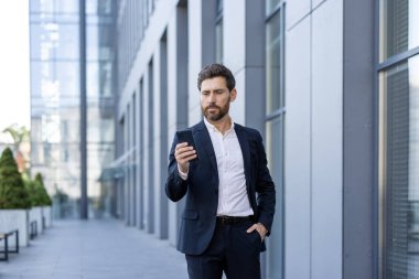 Takım elbiseli bir adam binanın dışında duruyor, telefonuna dalmış. Elini cebinde tutuyor, mesajlarını kontrol ederken ekrana odaklanıyor..