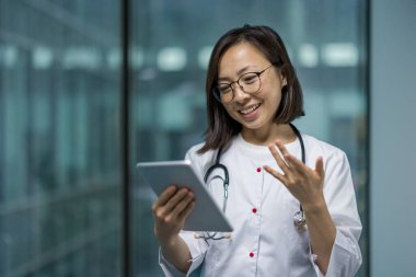 Genç başarılı bayan doktor video görüşmesi için tablet bilgisayar kullanıyor. Beyaz elbiseli kadın hastalara uzaktan danışıyor. Modern klinikteki Asyalı kadın..