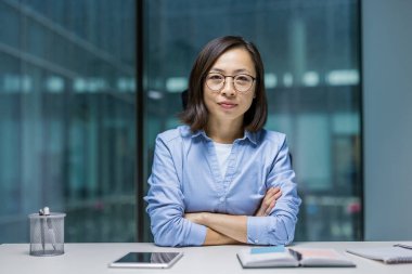 Gözlüklü, kendine güvenen Asyalı bir kadın kollarını kavuşturmuş, modern bir ofis ortamında çalışmaya hazır bir şekilde masada oturuyor.