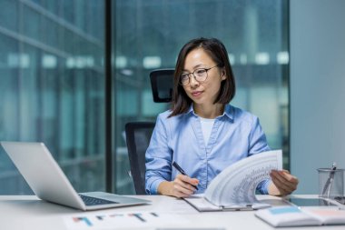 Ciddi bir şekilde evrak işleriyle uğraşan kendine güvenen bir iş kadını, başarılı bir finansör konsantre bir finansal rapor hazırlıyor. Ofisin içindeki masada çalışan Asyalı muhasebeci..