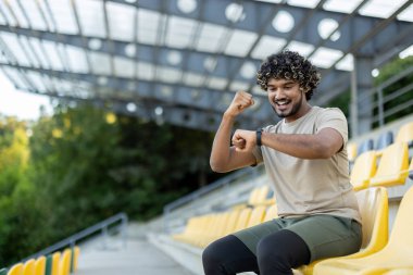 Genç sporcu başarılı antrenman sonuçlarını kutluyor, otururken dinleniyor. Adam akıllı saat fitness bilekliğinin verilerini kontrol ediyor, mutlu bir şekilde gülümsüyor, el ele tutuşuyor..