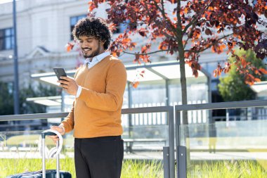 Genç adam elinde bavulla durakta toplu taşımayı bekliyor. Turist akıllı telefonundaki uygulamayı kullanarak mutlu bir şekilde gülümsüyor..