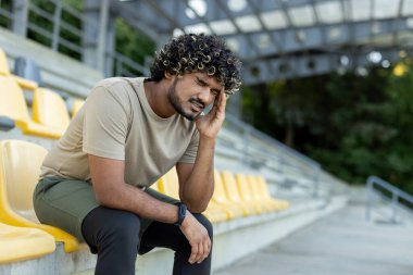Yorgun ve çalışkan bir sporcu, bir bankta oturuyor, aktif fiziksel egzersizler, fitness ve koşu sonrası dinleniyor. Baş ağrısından muzdarip.