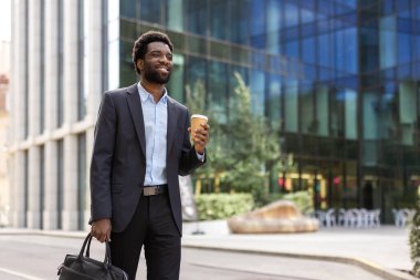 Genç ve başarılı bir işadamı elinde bir fincan sıcak içecek ve iş çantası çantasıyla ofis binasının önünde yürüyor. Takım elbiseli adam mutlu gülümsüyor..