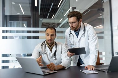 Beyaz önlüklü iki doktor bir dizüstü bilgisayarda, bir panoda ve modern ofis ortamında bir tablet üzerinde tartışıyorlar..