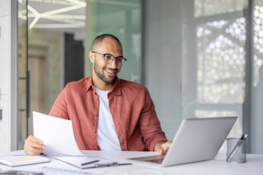 Gülümseyen bir adam, modern ofis ortamında dizüstü bilgisayarı üzerinde çalışırken belgelere göz atar. Uzaktan çalışma ya da çevrimiçi işbirliği önerir..