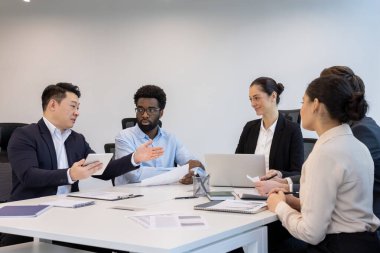 Bir grup iş adamı, bir toplantı için masa etrafında iş birliği yapıyor. Belgeleri gözden geçiriyorlar ve teknolojiyi kullanıyorlar..