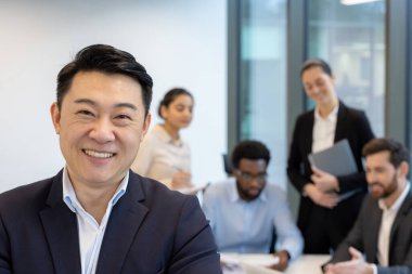 Asyalı bir işadamı ofisteki bir toplantıda ekibinin önünde kameraya güvenle gülümsüyor..