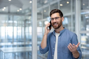 Mavi gömlekli bir işadamı telefon görüşmesi yapıyor. Modern bir ofis ortamında gülümsüyor ve el kol hareketi yapıyor..