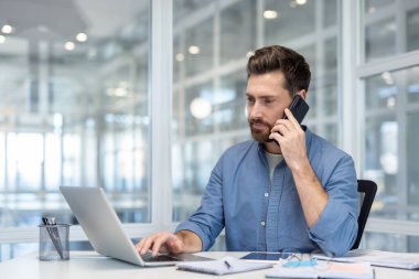 Odaklanmış bir iş adamı bilgisayarla çalışırken aynı anda akıllı telefonuyla modern ofis ortamında birden fazla iş yapar..