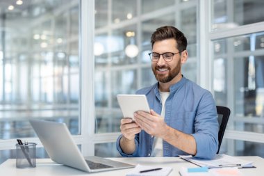 Bir adam modern ofis masasında tablet bir cihaz kullanırken gülümsüyor. Profesyonel biri meşgul görünüyor ve etrafı ofis ekipmanlarıyla çevrili..