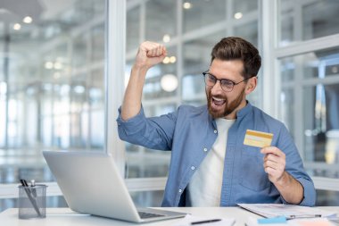 Bir adam online ödeme başarısını elinde bir kartla kutluyor, bir dizüstü bilgisayarın önünde oturuyor, mutlu..