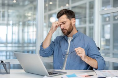 Stresli bir adam masasında oturmuş, işine ara verirken gözlerini ovuşturuyor..