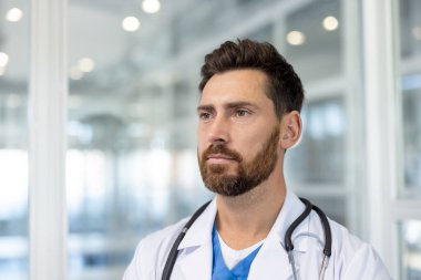 Düşünceli, olgun görünümlü bir doktor elinde steteskopla beyaz laboratuvar önlüğü giyerken görülüyor. Kararlı görünüyor..