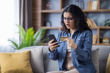 Endişeli genç Hintli bir kadın, elinde kredi kartıyla evdeki kanepede oturuyor ve şok içinde telefon ekranına bakıyor..