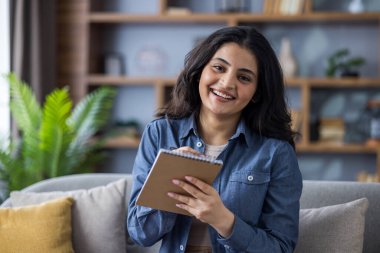 Evdeki gülümseyen Hintli genç bir kadının yakın plan portresi. Kameraya bakıyor ve not defterine not alıyor..