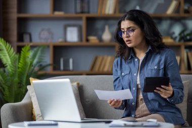 Endişeli genç Müslüman kadın evinde dizüstü bilgisayarın arkasında oturuyor, ekrana inançsızlıkla bakıyor ve elinde hesap makinesiyle elinde belgelerle bakıyor..