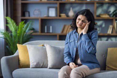 Müslüman genç bir kadın gömleğini giyip evdeki kanepede oturuyor, acı içinde yüzünü buruşturuyor, el ele tutuşuyor, baskıdan muzdarip..