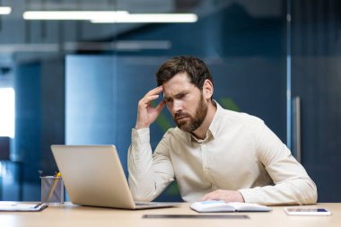 Ofiste oturan kendinden emin genç adam, başını tutuyor ve dizüstü bilgisayara endişeli bir şekilde bakıyor..