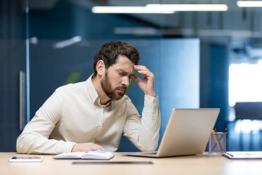 Ciddi ve yorgun genç bir iş adamı iş yerindeki ofiste dizüstü bilgisayarla oturur, gözlerini kapatır, başını eliyle tutar, şiddetli bir acı hisseder ve dinlenir..
