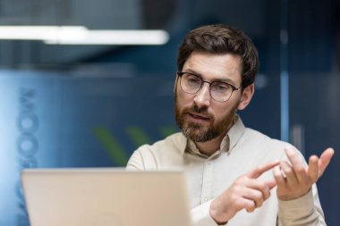 İş yerindeki ofiste dizüstü bilgisayarın önünde oturan genç bir iş adamının yakın plan fotoğrafı, bir video görüşmesinde konuşuyor ve kamerayı işaret ederken parmaklarını sayıyor..