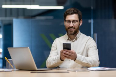 Genç bir ofis çalışanı iş yerinde oturuyor ve gülümseyerek cep telefonu kullanıyor..