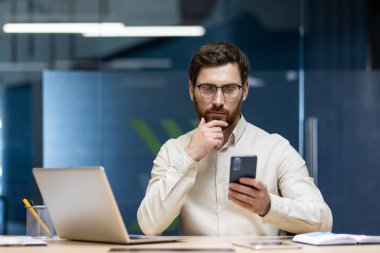 Ciddi bir genç adam ofiste çalışıyor, masada oturuyor, elini sakalına bastırıyor ve dikkatlice telefona bakıyor..