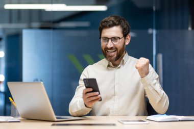 Genç ve mutlu bir adam ofiste oturuyor, gözlük takıyor, cep telefonuna gülümsüyor ve eliyle evet işareti yapıyor..