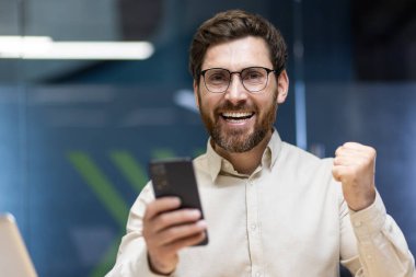 Gözlüklü mutlu bir gencin yakın plan portresi ve ofisteki açık renk gömlekli, başarı ve zaferle sevinen, elinde bir telefon tutan.