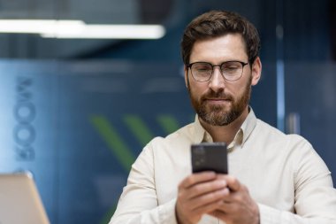 Ofiste olan ve elinde cep telefonu olan tişörtlü ve gözlüklü gülümseyen genç bir adamın yakın plan fotoğrafı..