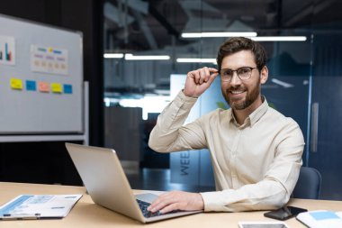 Genç, gülümseyen ve başarılı genç bir iş adamının portresi. Bir masada dizüstü bilgisayarla oturuyor, elinde gözlük ve kendinden emin bir şekilde kameraya bakıyor..
