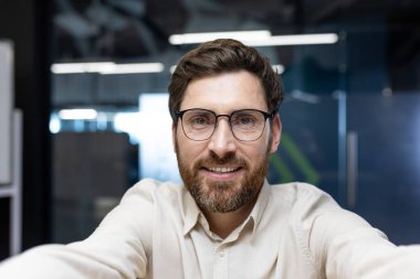 Gözlüklü ve sakallı genç bir adamın yakın plan portresi ofiste oturuyor, konuşuyor ve elinde tuttuğu kameraya bakıyor..