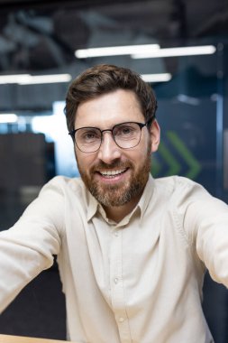 Ofiste elinde tuttuğu online kameraya dua eden gözlüklü ve gömlekli genç bir adamın dikey yakın plan fotoğrafı..