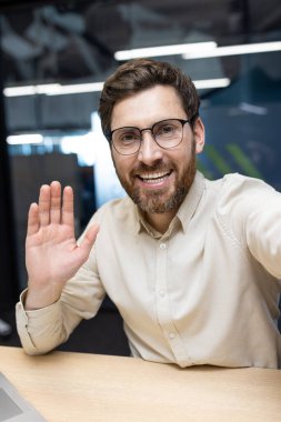 Ofisteki genç bir işadamının dikey yakın plan fotoğrafı, bir video görüşmesiyle konuşuyor, kameraya selam veriyor ve el sallıyor..