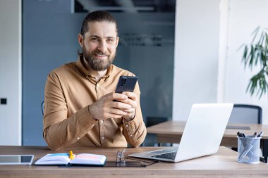 Sakallı, gülümseyen bir adam masasında odaklanmış ve meşgul bir şekilde otururken akıllı telefonunu kullanır. İşte açık dizüstü bilgisayar ipuçları.