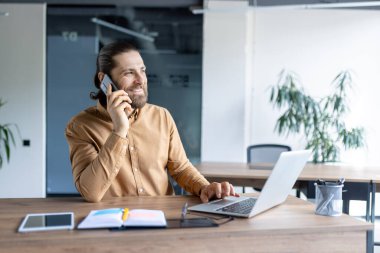 Ofiste akıllı telefondan konuşan bir adam gülümsüyor. Modern ofis ortamında masasında bir dizüstü bilgisayar üzerinde çalışıyor..