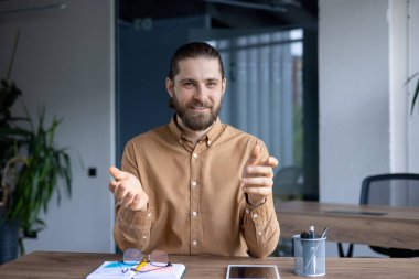 Video görüşmesi sırasında sakallı bir adam modern ofis ortamında ahşap bir masada oturuyor. Meşgul ve cana yakın görünüyor..