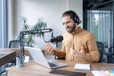 Sakallı bir adam mikrofon ve dizüstü bilgisayar kullanıyor. Bir ofis ortamında canlı yayın yapıyor..