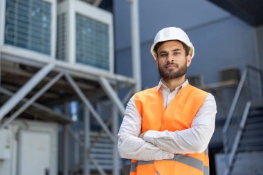 Sıkı bir şapka ve güvenlik yeleği giymiş bir inşaat işçisi kollarını kavuşturmuş, güvenli bir şekilde dışarıda duruyor. Ekipmanın yanında poz veriyor..