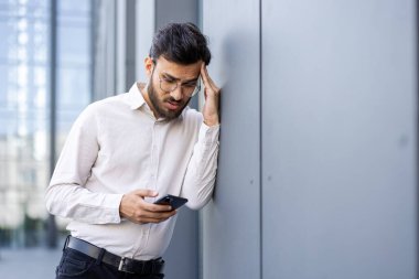 Gözlüklü endişeli bir işadamı dışarıda duvara yaslanırken telefonuna bakar. Gergin görünüyor..