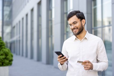 Gözlüklü çekici bir adam, modern bir binanın önünde kredi kartı ve akıllı telefonunu tutarken gülümsüyor..