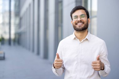 Neşeli bir işadamı, açık havada parlak bir gülümsemeyle, başarıyı ya da onayı kutlayarak baş parmak işareti yapar..