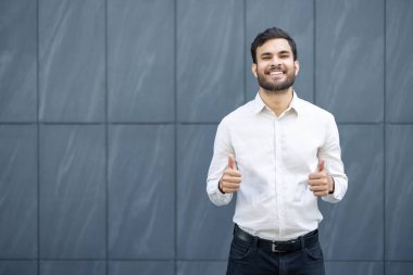 Siyah saçlı ve sakallı, gülümseyen bir adam beyaz gömlek giymiş, başparmak kaldırıyor. Arka plan gri desenli bir duvar..