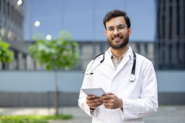 Gözlüklü gülümseyen bir doktor dışarıda steteskopu boynunda dururken bir tablet tutuyor..