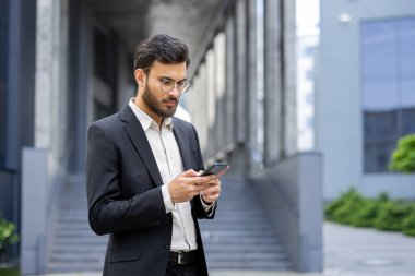 Takım elbiseli bir işadamı modern bir ofis binasının önünde dikilirken akıllı telefonuna dikkatle bakar. İçeriğe odaklanmış görünüyor..