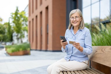Yaşlı bir kadın dışarıda oturuyor, telefonuna bakıp gülümsüyor ve elinde kredi kartı tutuyor. Güneşli bir günün tadını çıkarıyor..